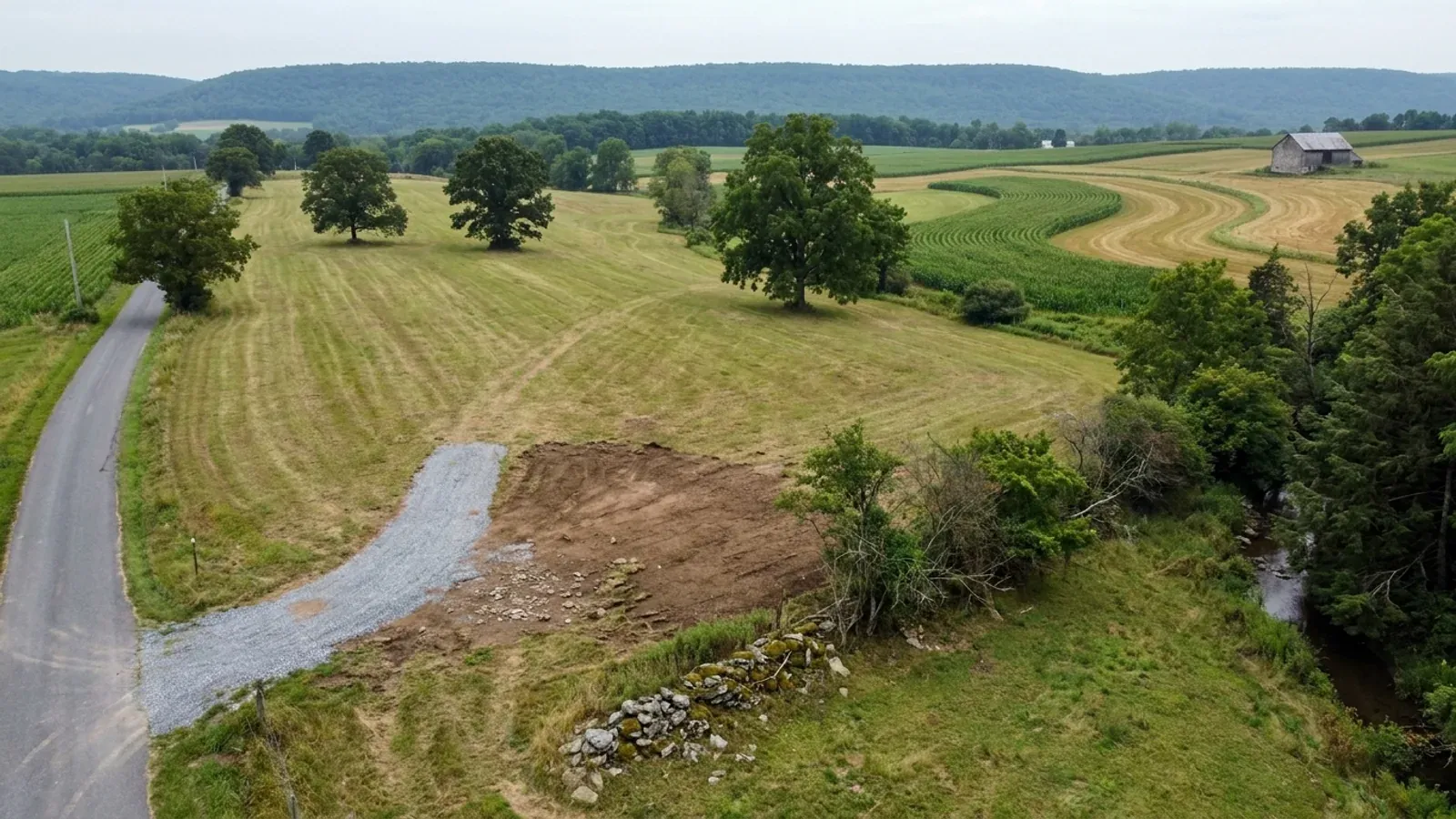Aerial view of an undeveloped rural lot at a hayfield edge with a stone lane, a hedgerow and contour-stripped farmland rolling toward a long wooded ridge.