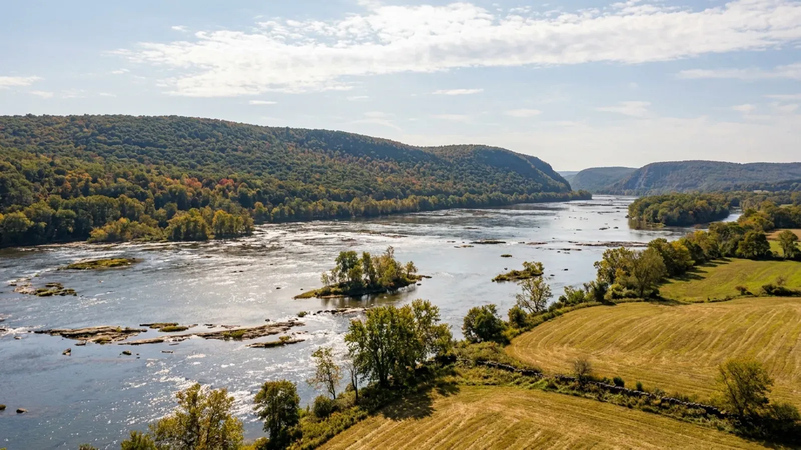 The wide, shallow Susquehanna River with rocky ledges and wooded islands below a long forested ridge, farmland on the near bank.